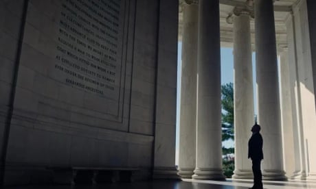 a man in shadow looks up at a plaque in a monument with white pillars