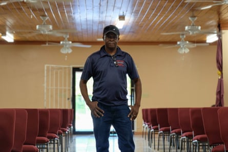 a man in a navy t-shirt and baseball cap stands in a room with ceiling fans and rows of red chairs