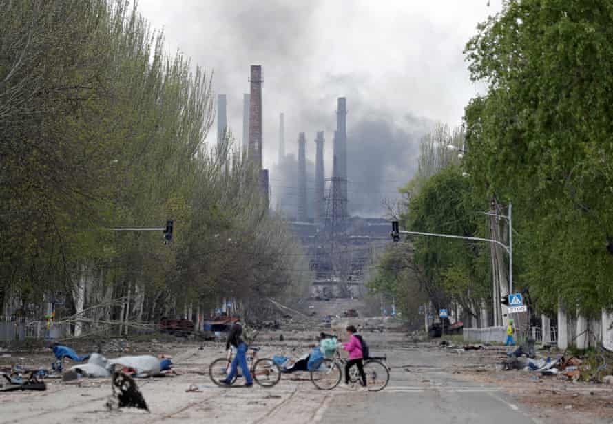 People walk their bikes across the street as smoke rises above a plant of Azovstal Iron and Steel Works in Mariupol, Ukraine.