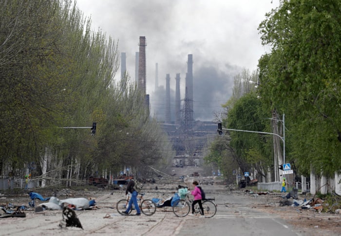 People walk their bikes across the street as smoke rises above the Azovstal steel plant in the southern port city of Mariupol.