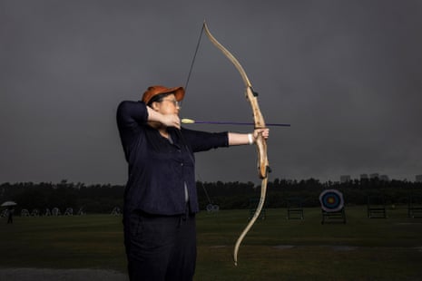A woman prepares to shoot an arrow from a large bow, in side profile. It is a dark and rainy day.