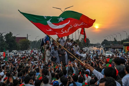 Someone waves a huge PTI flag among a crowd of protesters in a street.