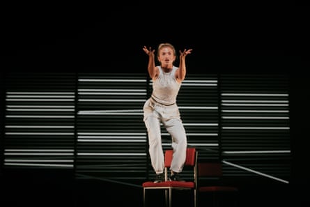 She stands on a gold chair with red upholstery, gesticulating forwards with both hands