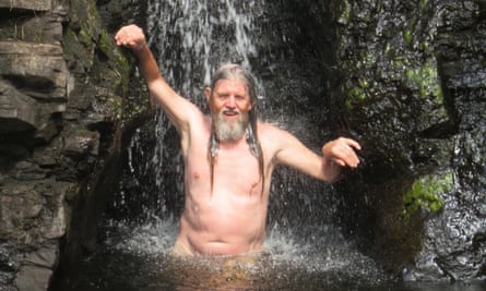 John King stands naked in water with a waterfall flowing on to his long hair.
