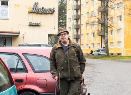 Pentti Korhonen stands by a red car using a cane, with apartment blocks behind him in Otanmäki.