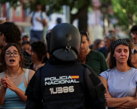 A closeup, back-facing shot of a Spanish National Police officer (POLICIA NACIONAL) wearing a black helmet and tactical riot gear, facing a crowd of emotional housing activists