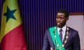 Bassirou Diomaye Faye speaks at a podium wearing a green sash and ceremonial chains of office with a Senegalese flag in the background