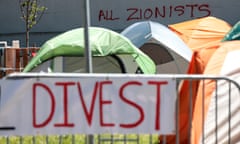 A lawn full of tents, with a handmade sign in the foreground that says Divest, and red spray paint on a wall beyond the tents that says All Zionists.