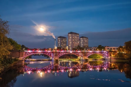 A night view of a bridge with illuminated city towers, a distillery chimney and colourful lights reflected in the river