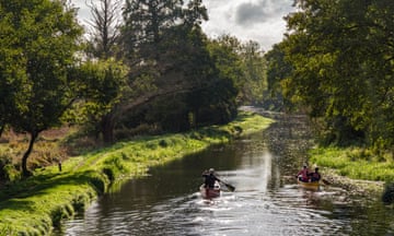 Three people paddle down a river in two canoes with trees on either side