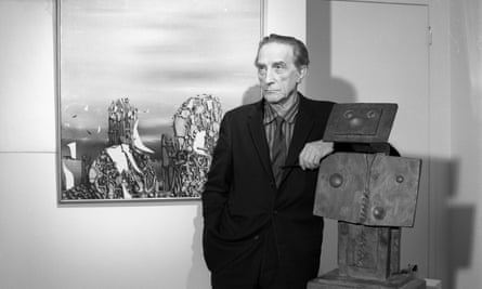 Artist Marcel Duchamp in 1960. He leans on a sculpture and stands in front of an abstract paining. He looks into the distance.