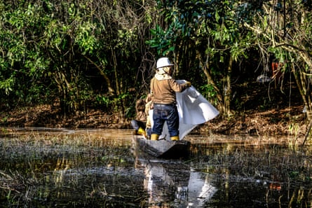 Three men are in a small boat, with one in a hard hat standing and holding a white sheet out over oily water