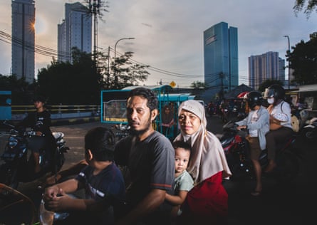 71% 28°C A family rides a scooter in the Kemayoran neighborhood in the city center of Jakarta, with high-rise middle-class buildings in the background. Indonesia, 2022