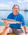 A man in a blue jumper sits in the sunshine on a harbour wall hoolding a fish.