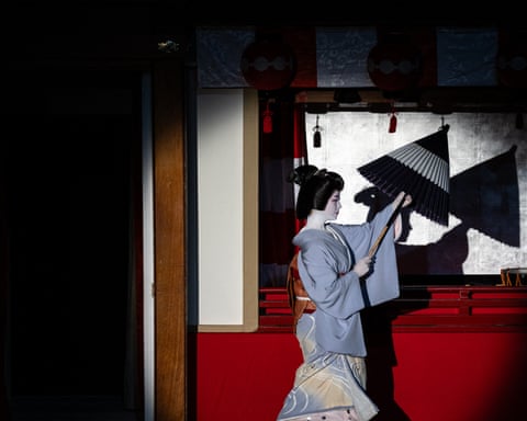 A woman with a painted white face and a blue kimono holds a black parasol