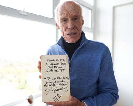 An elderly white-haired man stands by a window holding a rectangular item with a message and signature on it