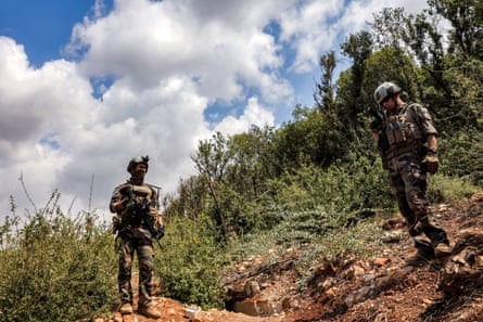 Two soldiers in camouflage fatigues and helmets, carrying guns, stand on a rocky hillside with bushes behind them; the sky is blue with white clouds