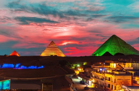 Three pyramids illuminated in red, yellow and green stand against a vivid sunset, with Cairo’s buildings lit up in the foreground.