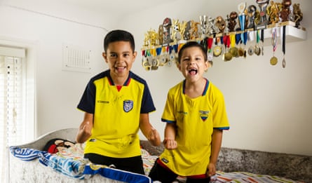 Juan Alejandro Pachar Carmona and Juan Esteban Pachar Carmona, wearing the Equador World Cup kit sitting on a top bunk bed at home with lots of trophies and medals on a shelf behind them