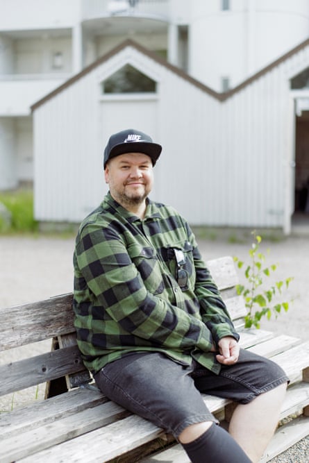 A smiling man with stubble and wearing a baseball cap sitting outside on a bench