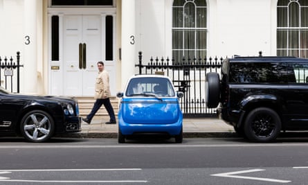 The Microlino parked between two larger vehicles with a white house behind