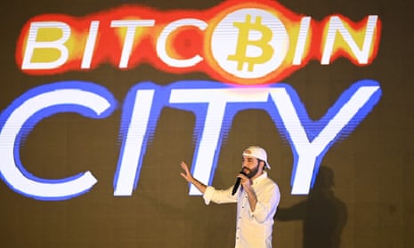 Man wearing white shirt and backward cap gesturing in front of a sign that reads 'Bitcoin City'.