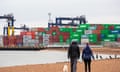Shipping containers are unloaded from a cargo ship at the Port of Felixstowe in Suffolk as two people walk a dog on a beach