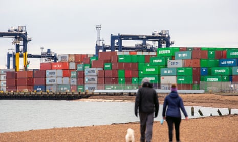 Shipping containers are unloaded from a cargo ship at the Port of Felixstowe in Suffolk as two people walk a dog on a beach