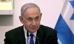 A man in a black suit and blue tie speaks into a microphone, with an Israeli flag hanging behind him