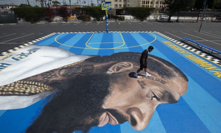 A man walks on a basketball court mural of a profile view of Nipsey Hussle on a blue striped background.