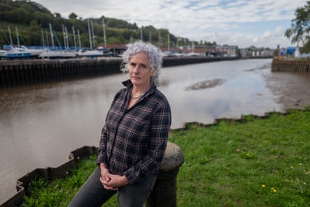 Olivia Doherty stands on grass by the riverside with moored boats in the background on the other bank. She has curly grey hair and wears a checked shirt and jeans.