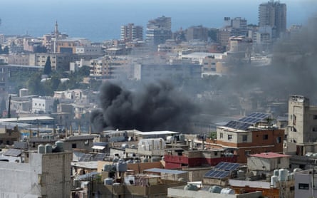 Black smoke billows from a dense cityscape with the sea in the background