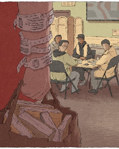 A group of four men sit around a small folding table inside a room with beige walls and carpeted flooring. A man with many hospital tags stands in the foreground. responsive-image