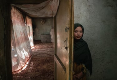 Amina, an Afghan girl in a green headscarf, peers from behind a wooden door into a dim room
