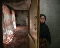 Amina, an Afghan girl in a green headscarf, peers from behind a wooden door into a dim room