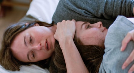 two young women with long brown hair lie together in an embrace; they look as if they are or have been crying