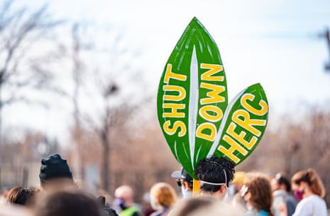 A large leaf-shaped sign reading "Shut Down HERC" is held above a crowd at a Minneapolis protest
