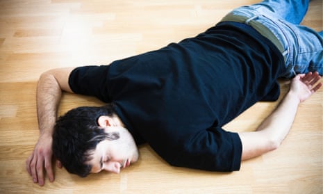 Darked haired man in dark-blue T-shirt and jeans lying, eyes closed, face down on a wooden floor.