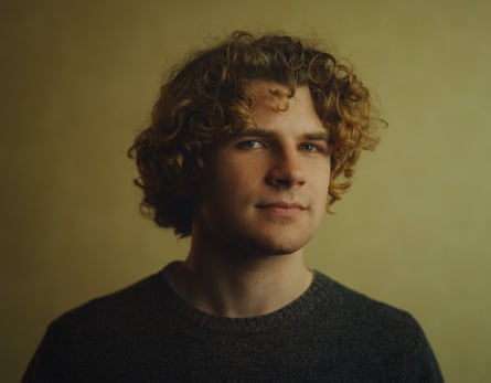 A man with curly hair and a black top, a yellow background is behind him