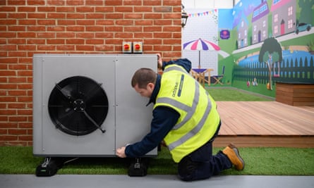 An engineer in a hi-vis vest kneels by a heat pump, which looks similar to an air conditioning unit, on an outside wall