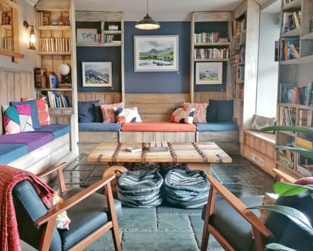 A lounge room with a coffee table, benches, armchairs, beanbags and cushions, with books on shelves and paintings on the wall