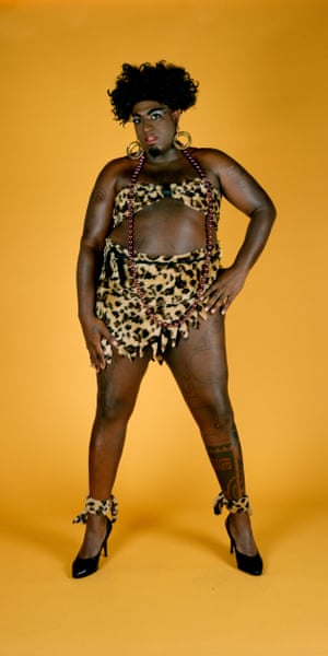 A drag performer wearing leopard print bra and matching skirt and high heels in front of a yellow-orange backdrop