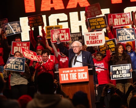 Senator Bernie Sanders raises a manus successful acknowledgment while speaking astatine a plinth pinch a ‘tax nan rich’ banner connected it