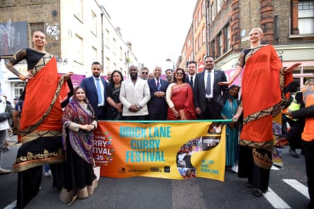 People - including two women on stilts - gather around a banner saying ‘Brick Lane curry festival’