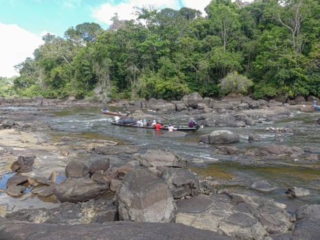 Two pirogues are steered, poled and pulled along a channel through rocks on a wide river bordered by jungle