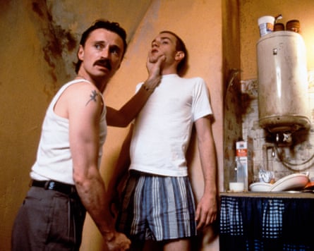 Two men in white singlets and a T-shirt, one with his back against the wall, the other holding his throat. The background is a squalid kitchen