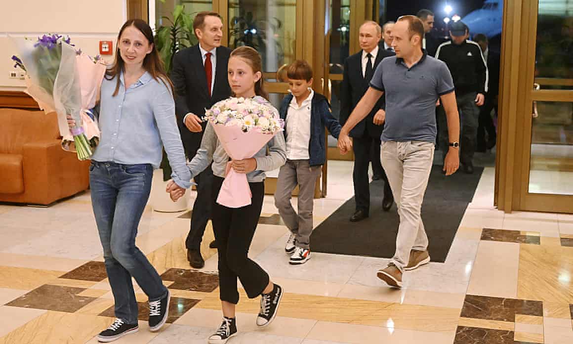 Vladimir Putin walks behind a smiling Anna Dultseva, who is walking hand-in-hand with her daughter and holding large bouquets of flowers, trailed by Artyom Dultsev holding the hand of his son