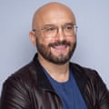 A man with a bald head, a beard and glasses, wearing a blue jumper and leather jacket