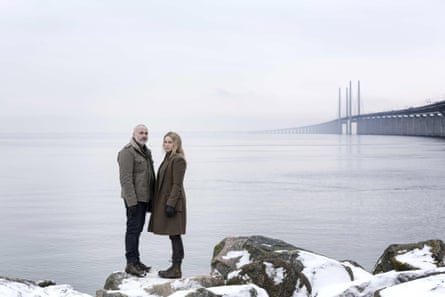 Two lead actors from The Bridge standing on rocks near sea with bridge in the background
