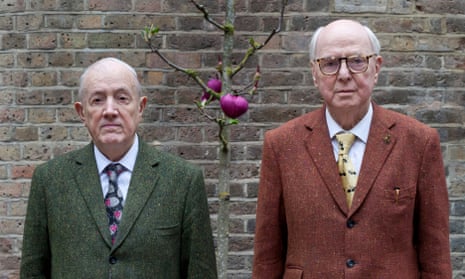 Gilbert, in a green tweed suit, and George, in an orange tweed suit, pose for a photograph on either side of a small bare tree with pink hearts tied to it